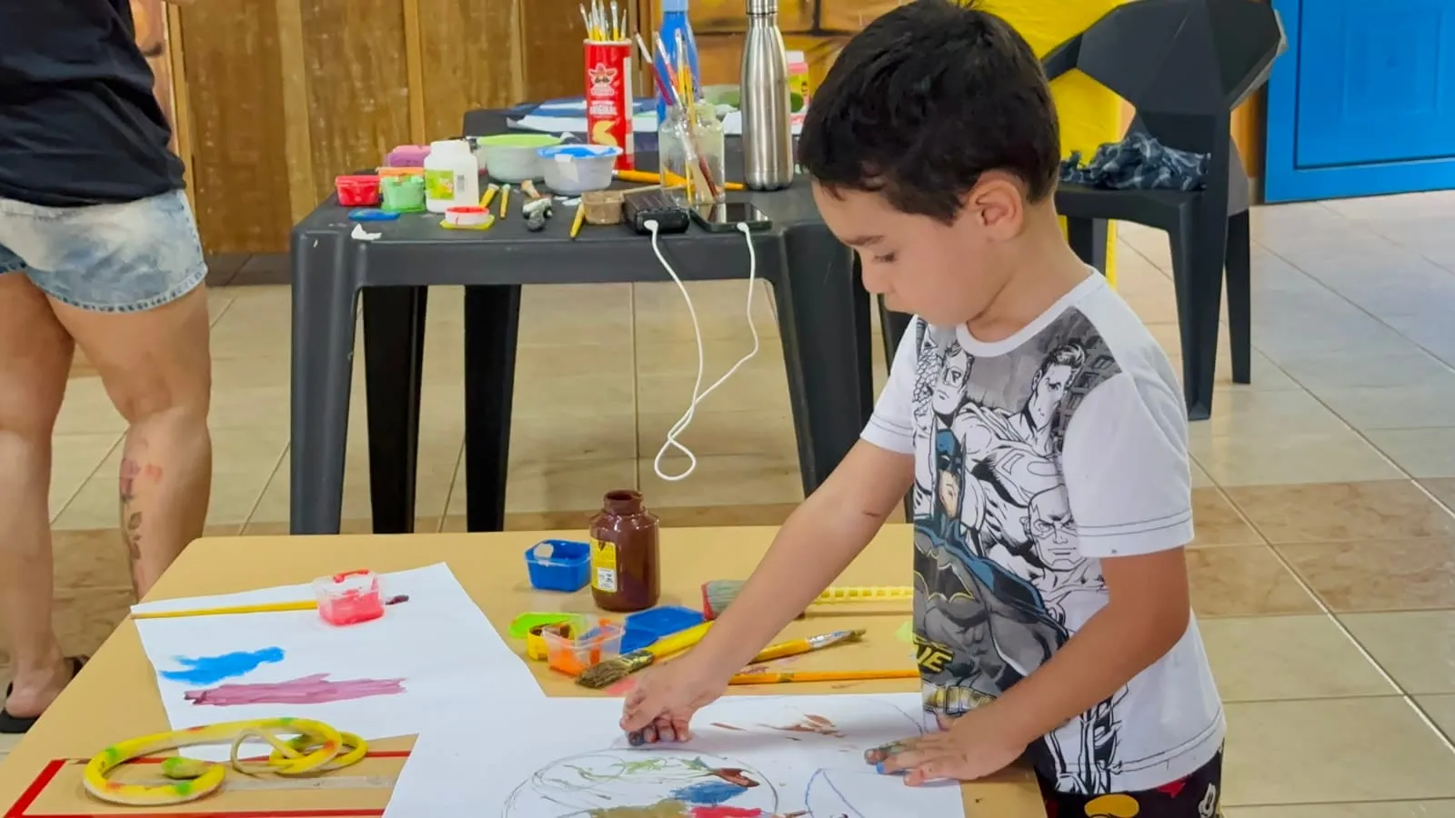 Menino desenhando e pintando no papel em uma mesa com potes de tinta