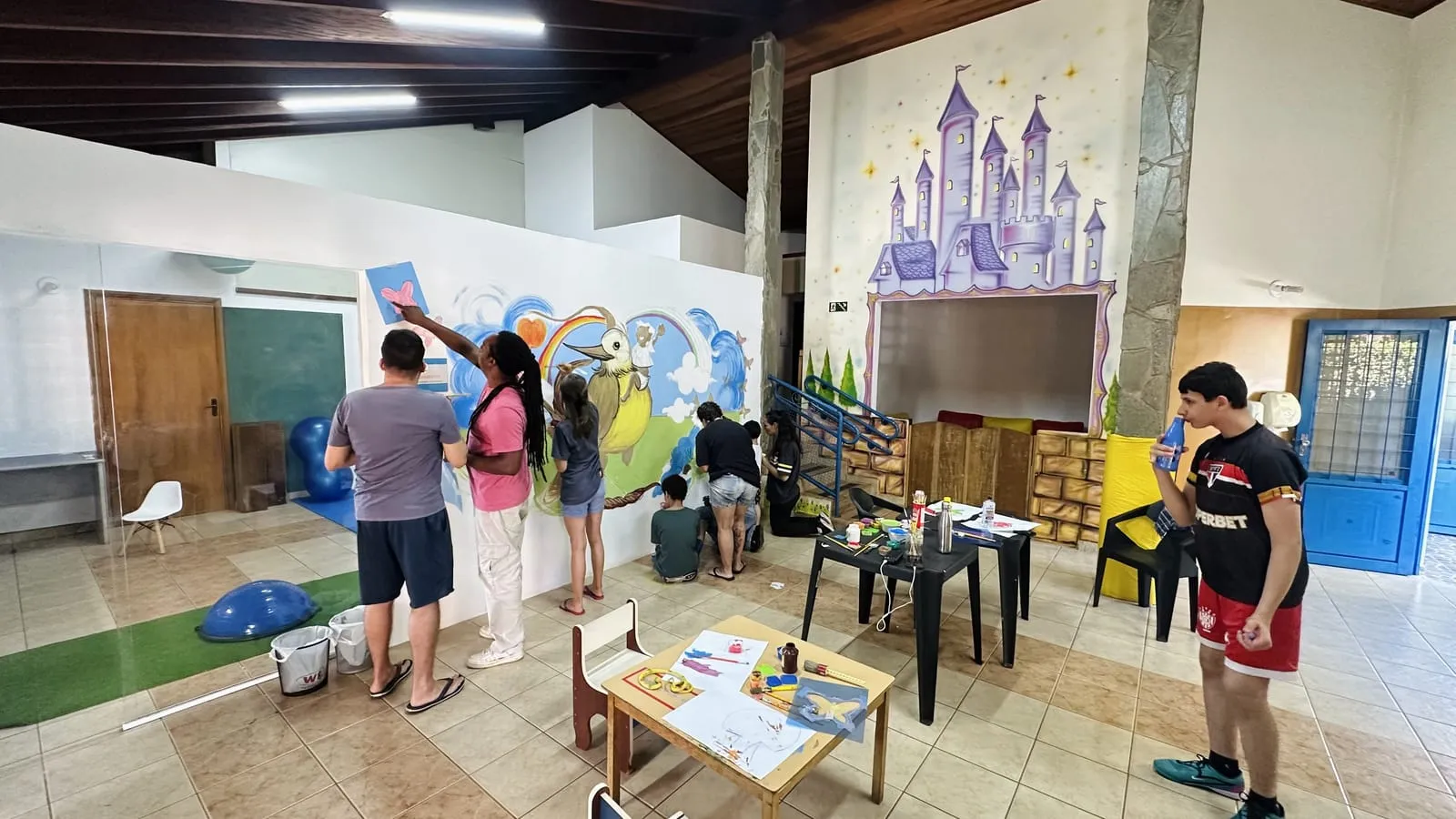 Salão da Te Apoio com o mural em pintura, participantes pintando a parede e mesas com tintas